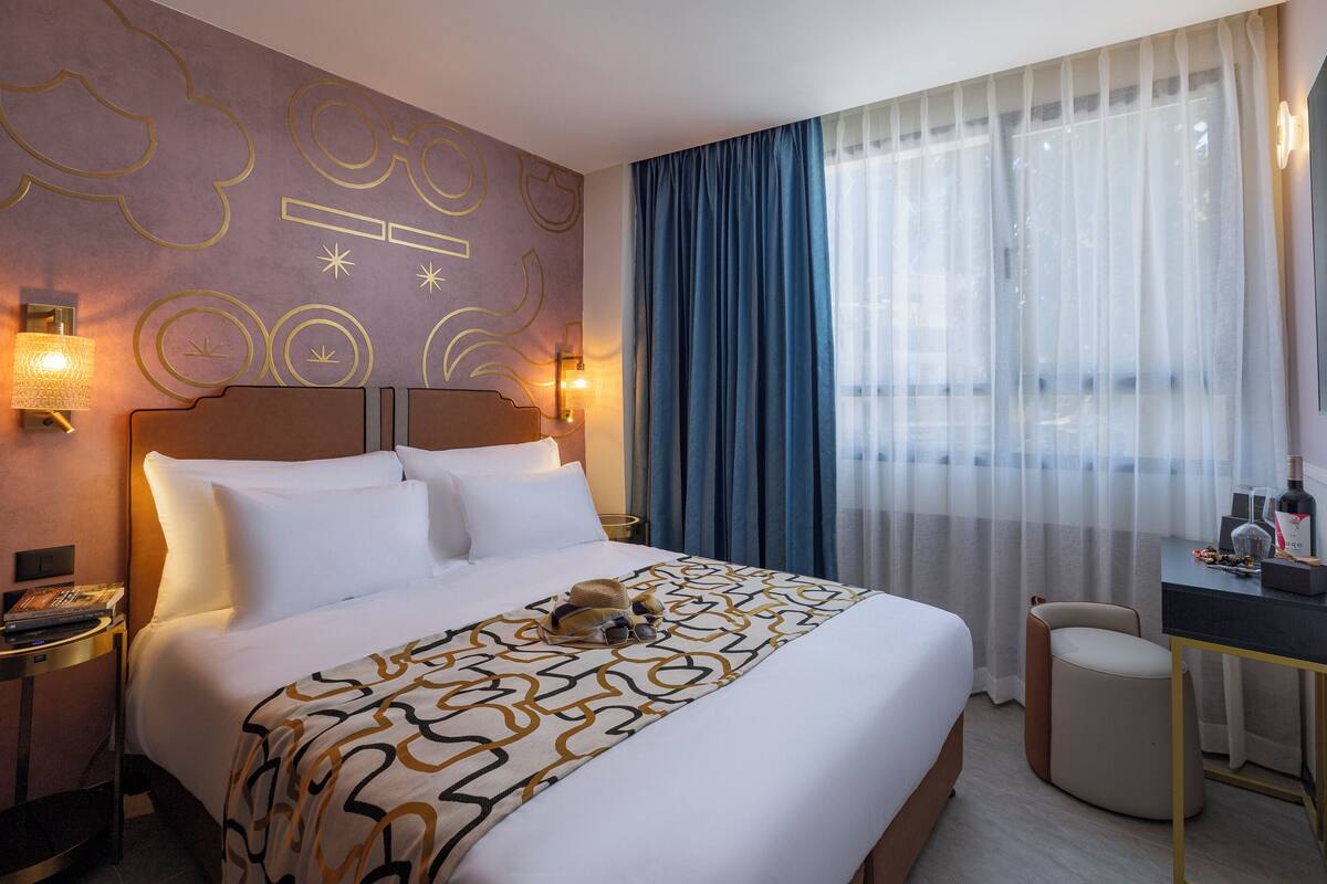 Una habitación de hotel con una cama grande, un escritorio con una silla, una mesita de noche e iluminación decorativa en la pared.