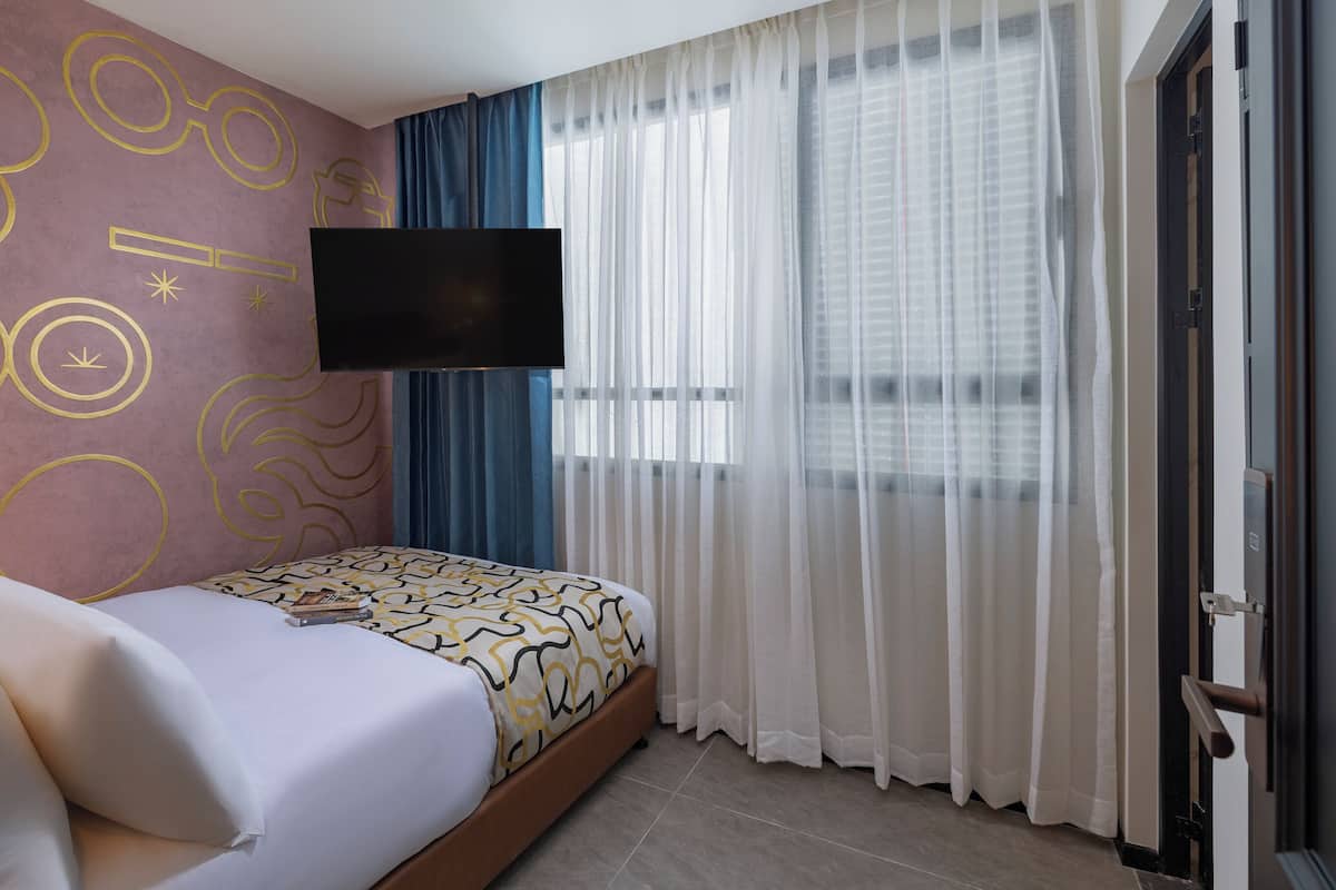 Habitación de hotel con cama, televisor de pantalla plana en la pared y una ventana grande con cortinas traslúcidas.