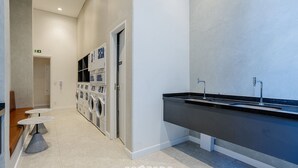 A modern bathroom with a black sink, a mirror, and a wall-mounted toilet. There are two small round tables with chairs, and a door leading to a laundry area with washing machines. - Modern and Fully Equipped Apartments, With Leisure Facilities and in the Best Location in Pinheiros - SP - EGP (São Paulo)