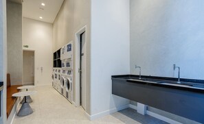 A modern bathroom with a black sink, a mirror, and a wall-mounted toilet. There are two small round tables with chairs, and a door leading to a laundry area with washing machines.