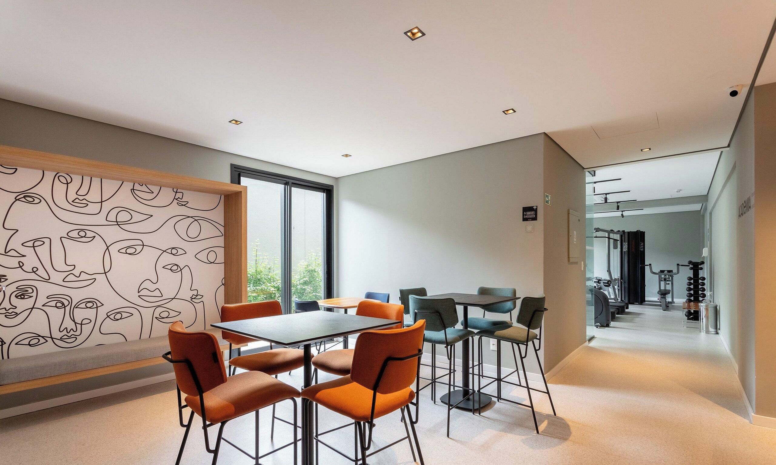 Un intérieur moderne avec un coin repas comprenant des chaises et une table orange, un mur orné d’œuvres d’art abstraites en forme de lignes, et une vue sur une salle de sport en arrière-plan.