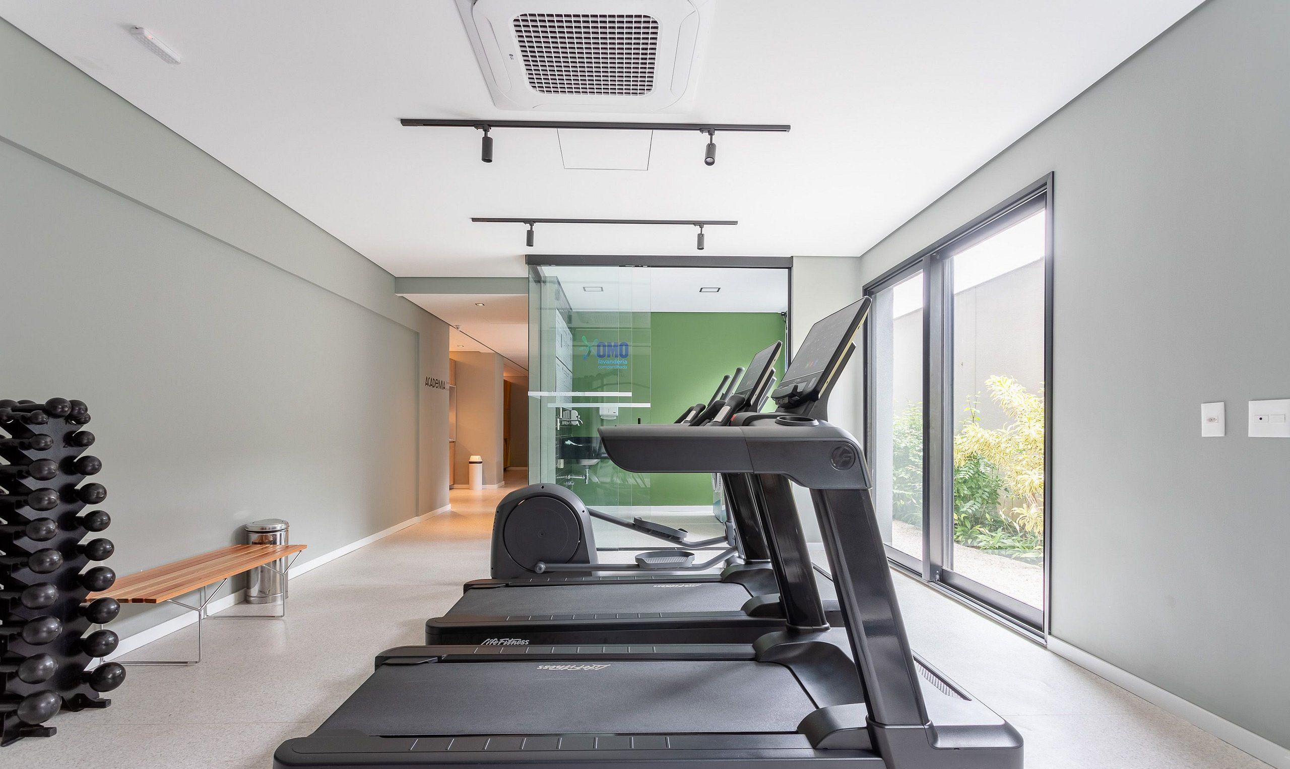 Une salle de sport moderne équipée de tapis de course, d’un porte-haltères fixé au mur et d’une grande fenêtre laissant entrer la lumière naturelle.