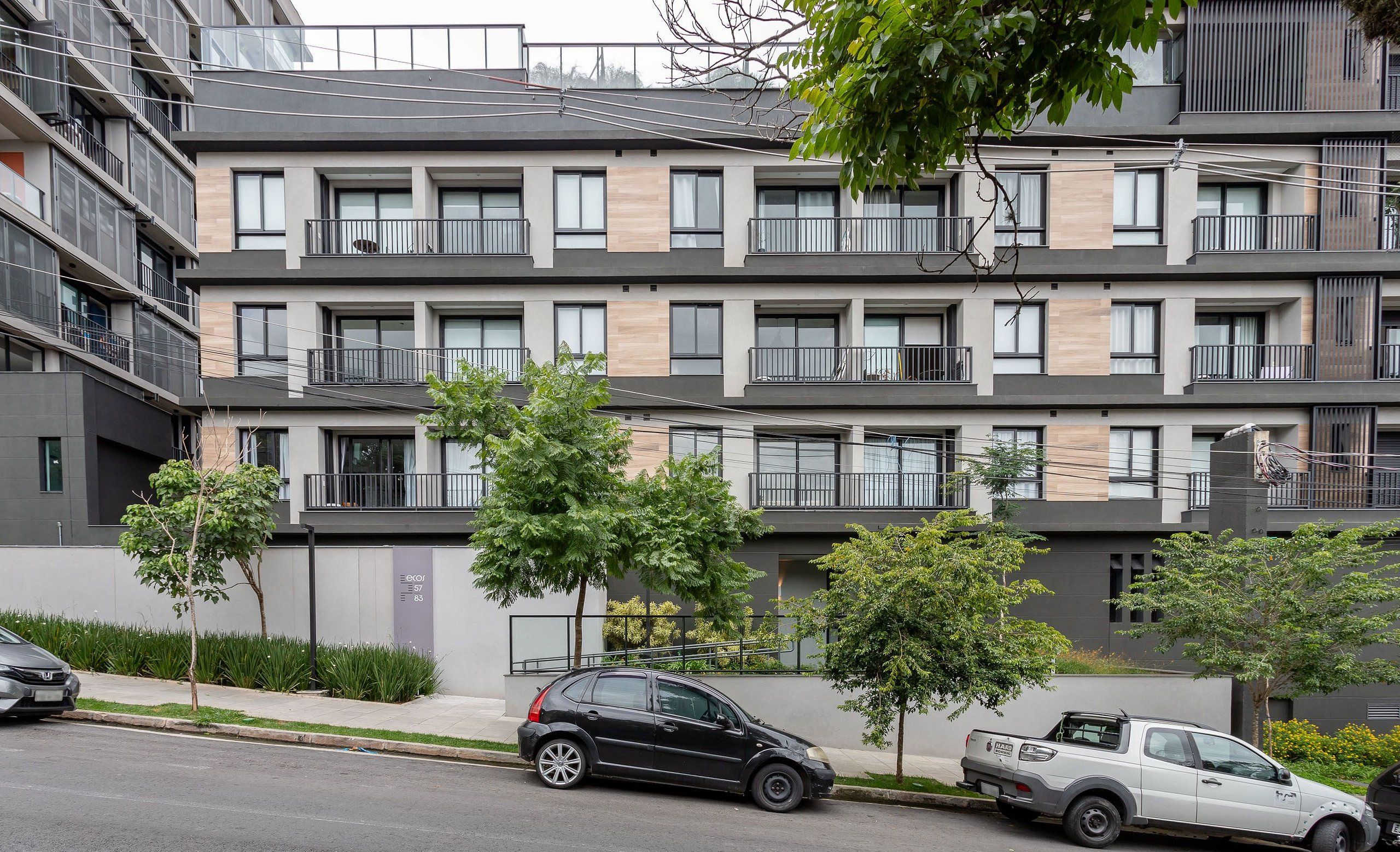 Un immeuble résidentiel à plusieurs étages, doté de balcons, d’un espace de stationnement et d’un petit arbre au premier plan.