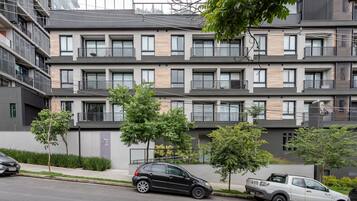 Un immeuble résidentiel à plusieurs étages, doté de balcons, d’un espace de stationnement et d’un petit arbre au premier plan.