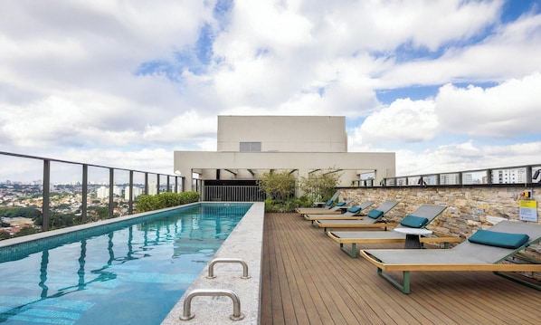 A rooftop pool with lounge chairs, a stone wall, and a view of the city. - Estúdio em SP perto do metrô | GBA908 (São Paulo)