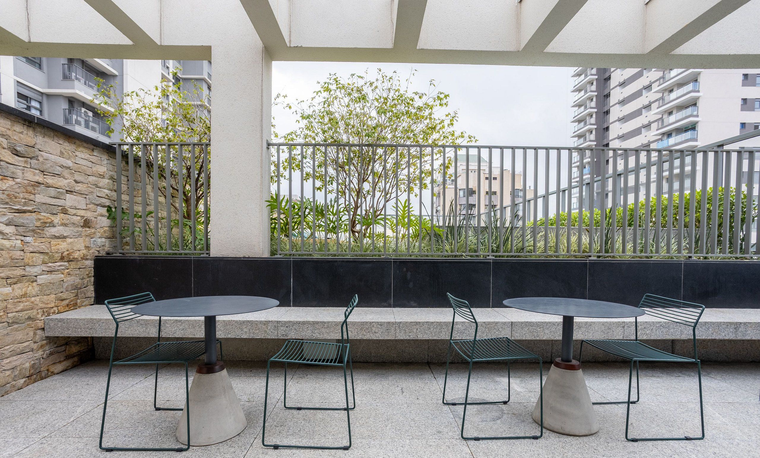 An outdoor patio with two round tables and four chairs, surrounded by a stone wall and metal railing, overlooking a residential area.