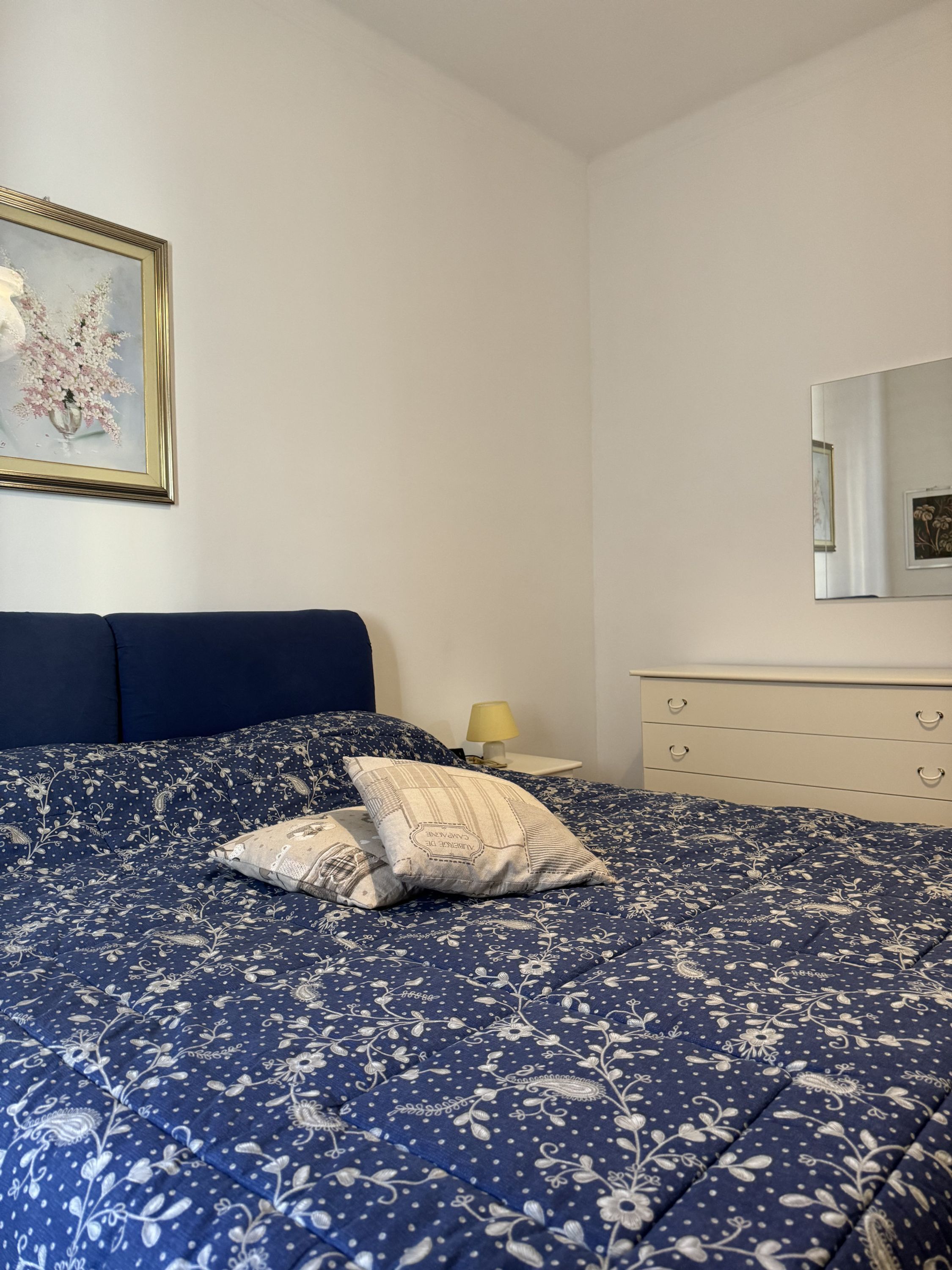 Uma cama bem arrumada com um edredom estampado com flores, uma mesa de cabeceira com abajur e uma obra de arte floral emoldurada na parede.