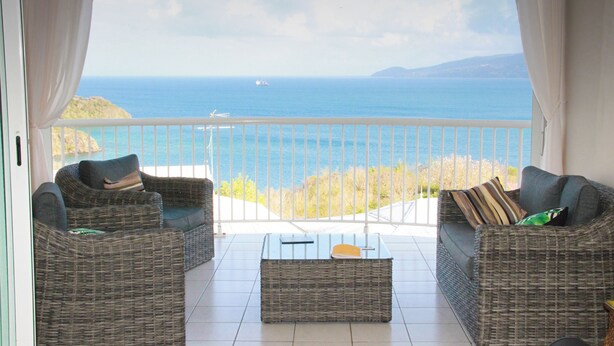 A balcony with wicker furniture, a glass coffee table, and a view of the sea and distant hills. - Superb Caiali Blue apartment (Les Trois-Îlets)