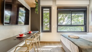 A modern room with a bed, a dining table with chairs, a TV mounted on the wall, and a window with a view of trees. - Beautiful Studio With Pool in Campo Belo Hmc0320 (São Paulo)