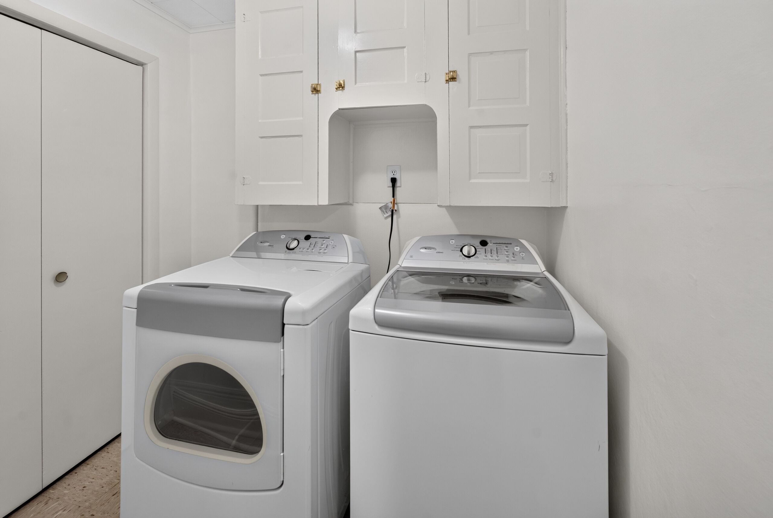 Une buanderie équipée de deux machines à laver à chargement frontal et d’un s èche-linge, avec des placards blancs au-dessus et une porte sur le côté gauche.