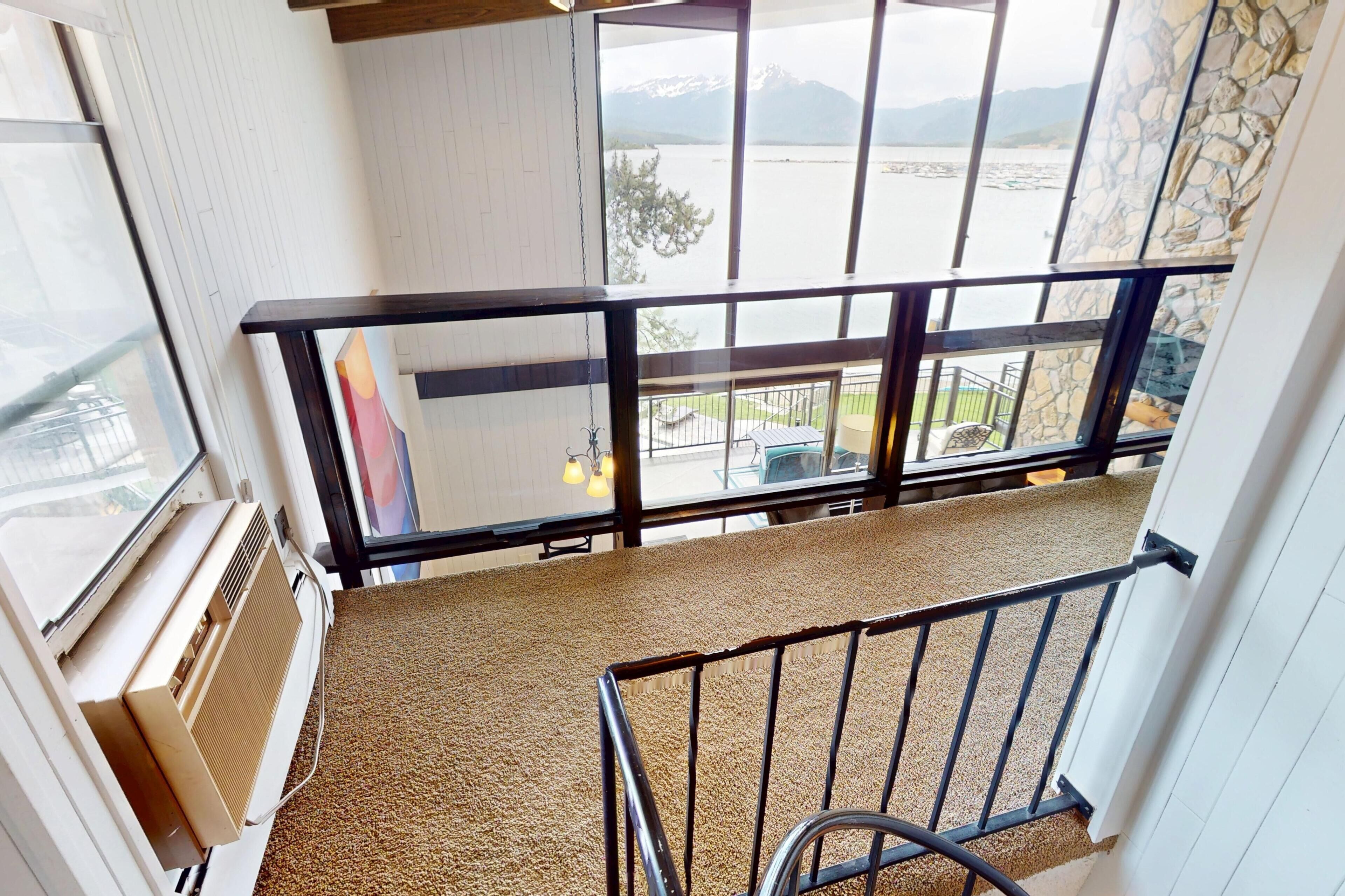 A balcony with a view of a lake and mountains, a metal railing, and a carpeted floor.