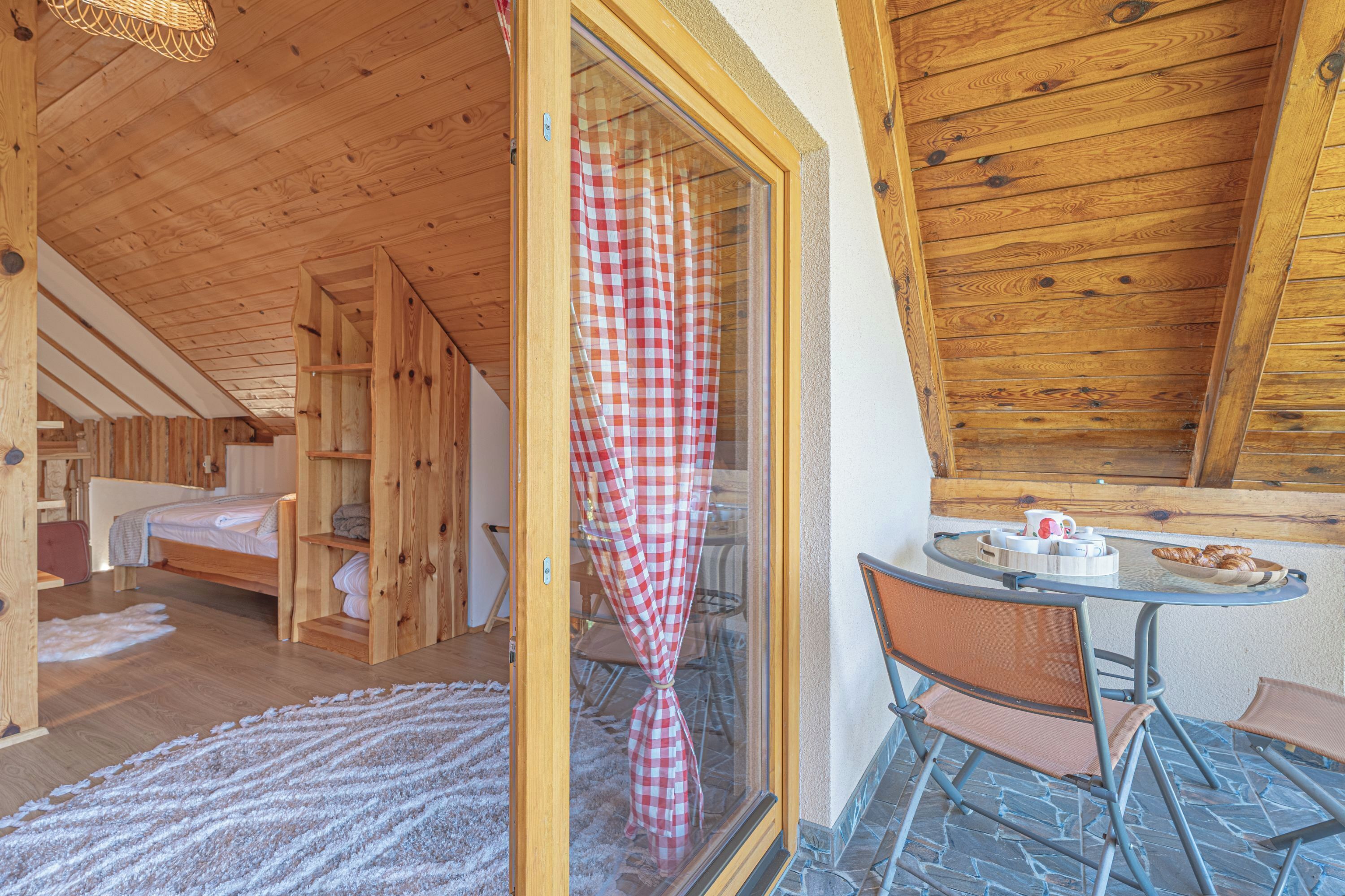 A wooden cabin interior with a dining area, a bed, and a window with a checkered curtain.