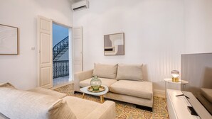 A modern living room with a beige sofa, a coffee table, and a TV stand. There is a staircase leading to an upper floor and a door to the left. - Impressive 3 bedrooms, sun terrace, jacuzzi AMRA1-1 (Sliema)