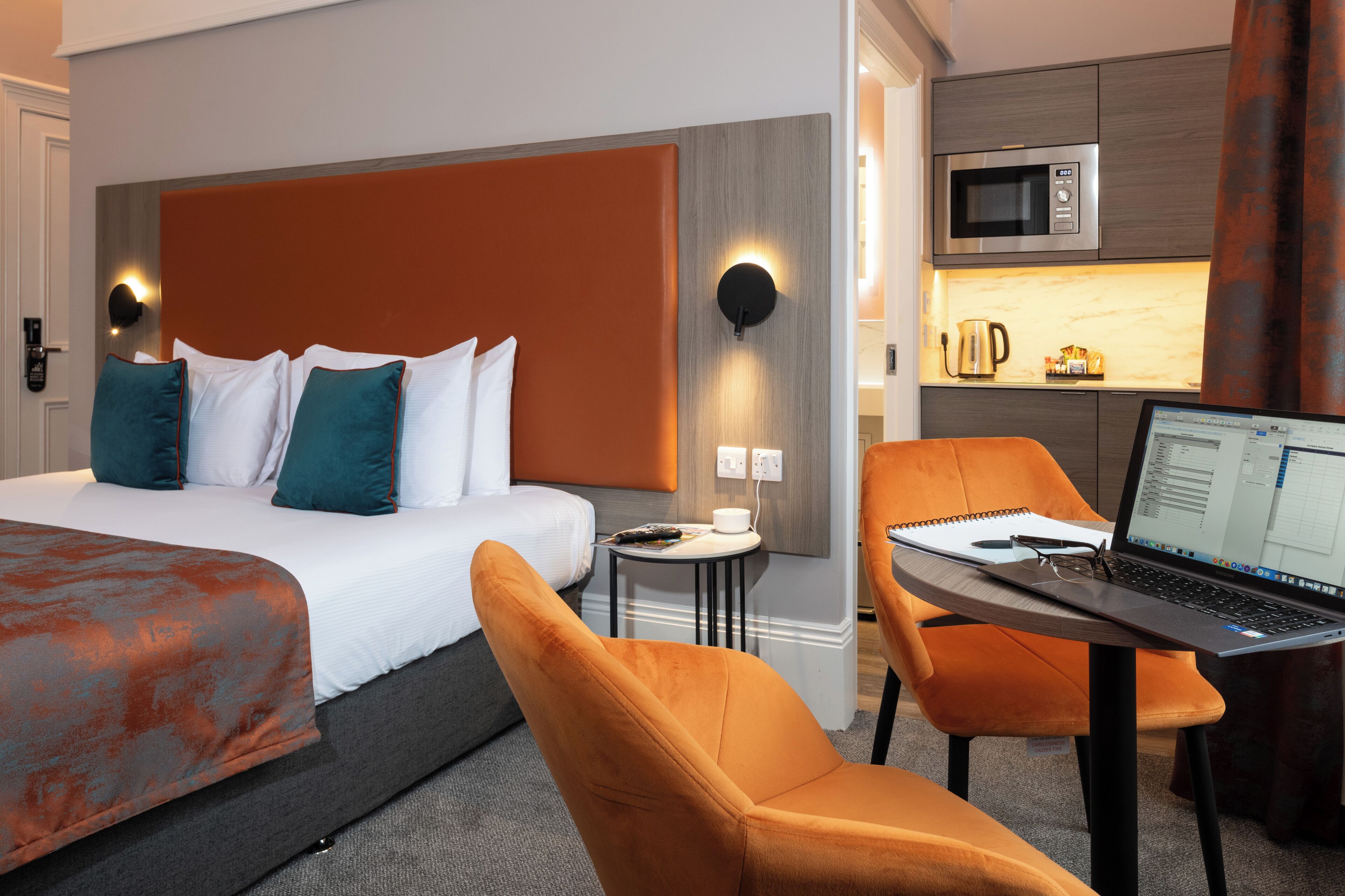 Une chambre d’hôtel avec un lit, un bureau avec un ordinateur portable, un four à micro-ondes et une petite table avec une tasse et une télécommande.