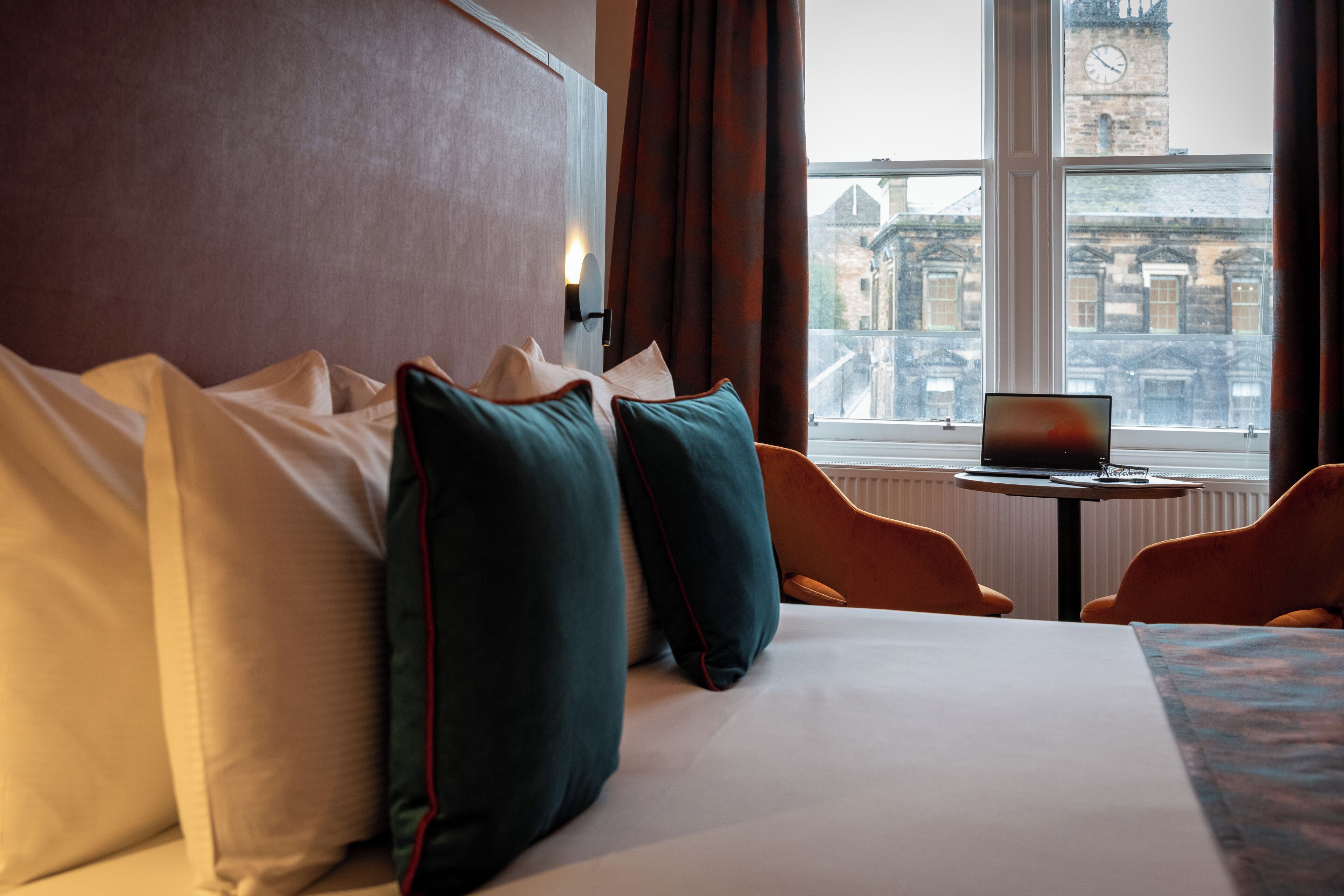 A hotel room with a bed, pillows, a laptop on a small table, and a view of a historic building through a window.