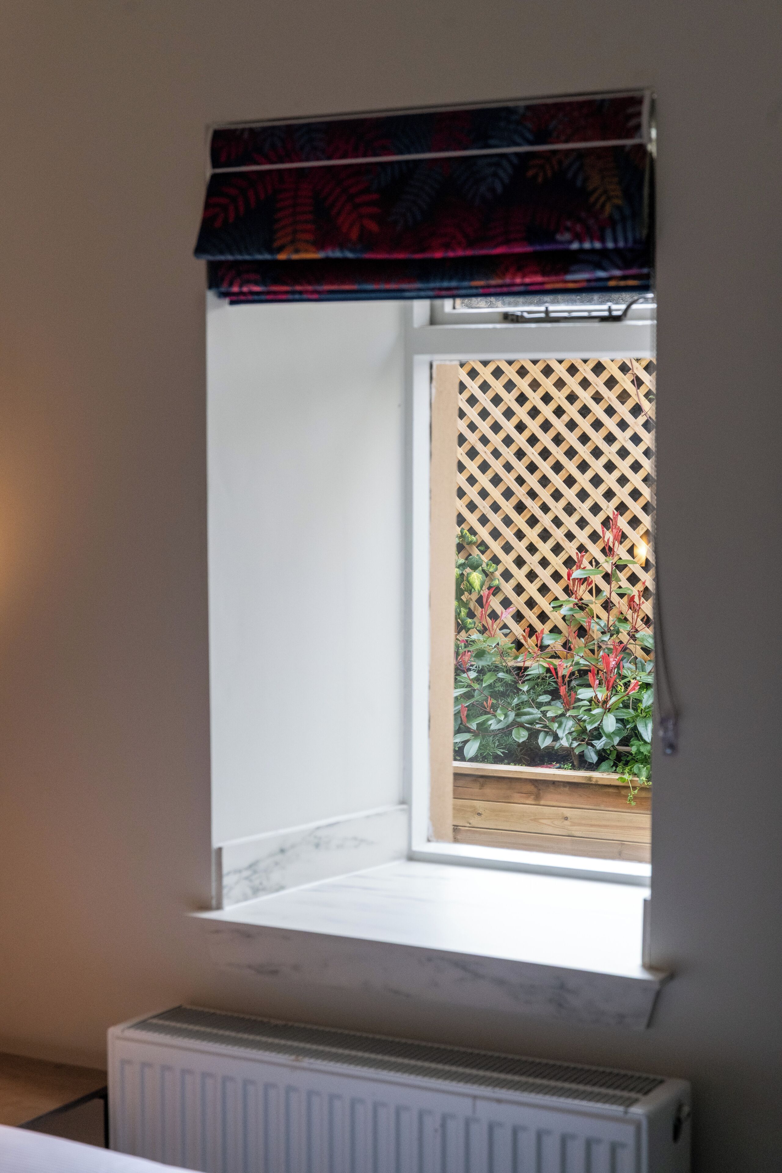 A window with a wooden lattice screen, a radiator below, and a view of a garden with red-leafed plants.
