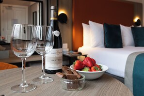 A hotel room with a bed, a bedside table with wine glasses, a bottle of wine, and a bowl of strawberries.