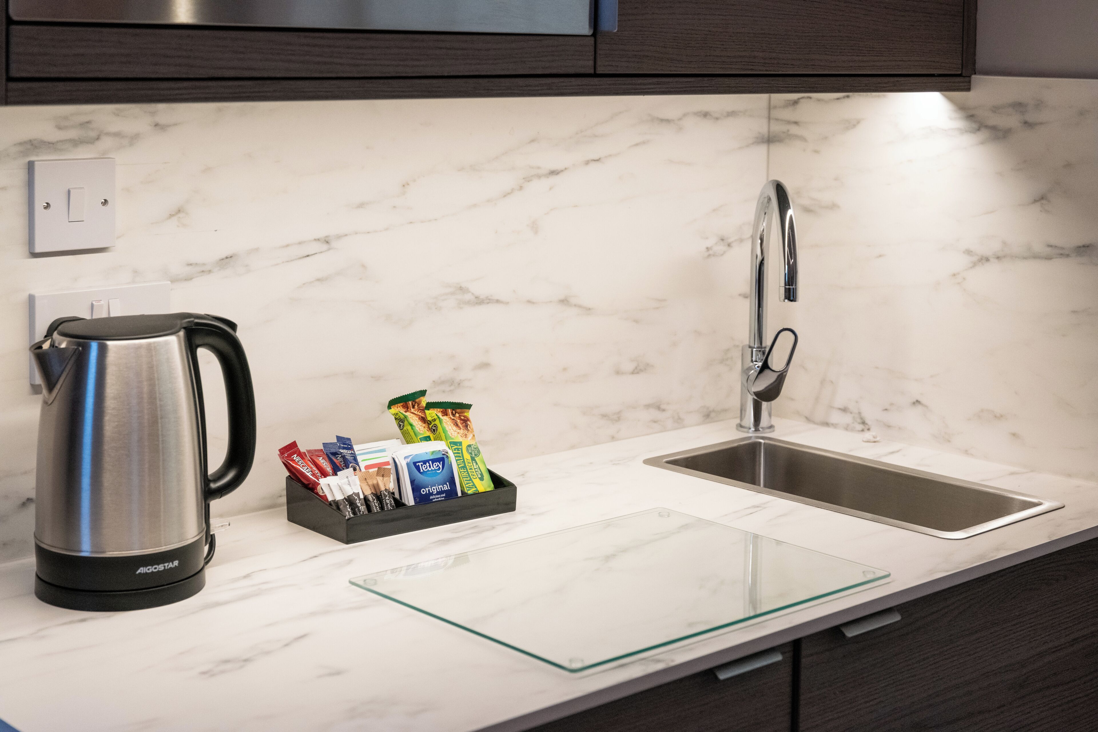 A kitchen with a stainless steel kettle, a glass container with snacks, a modern faucet, and a marble countertop.