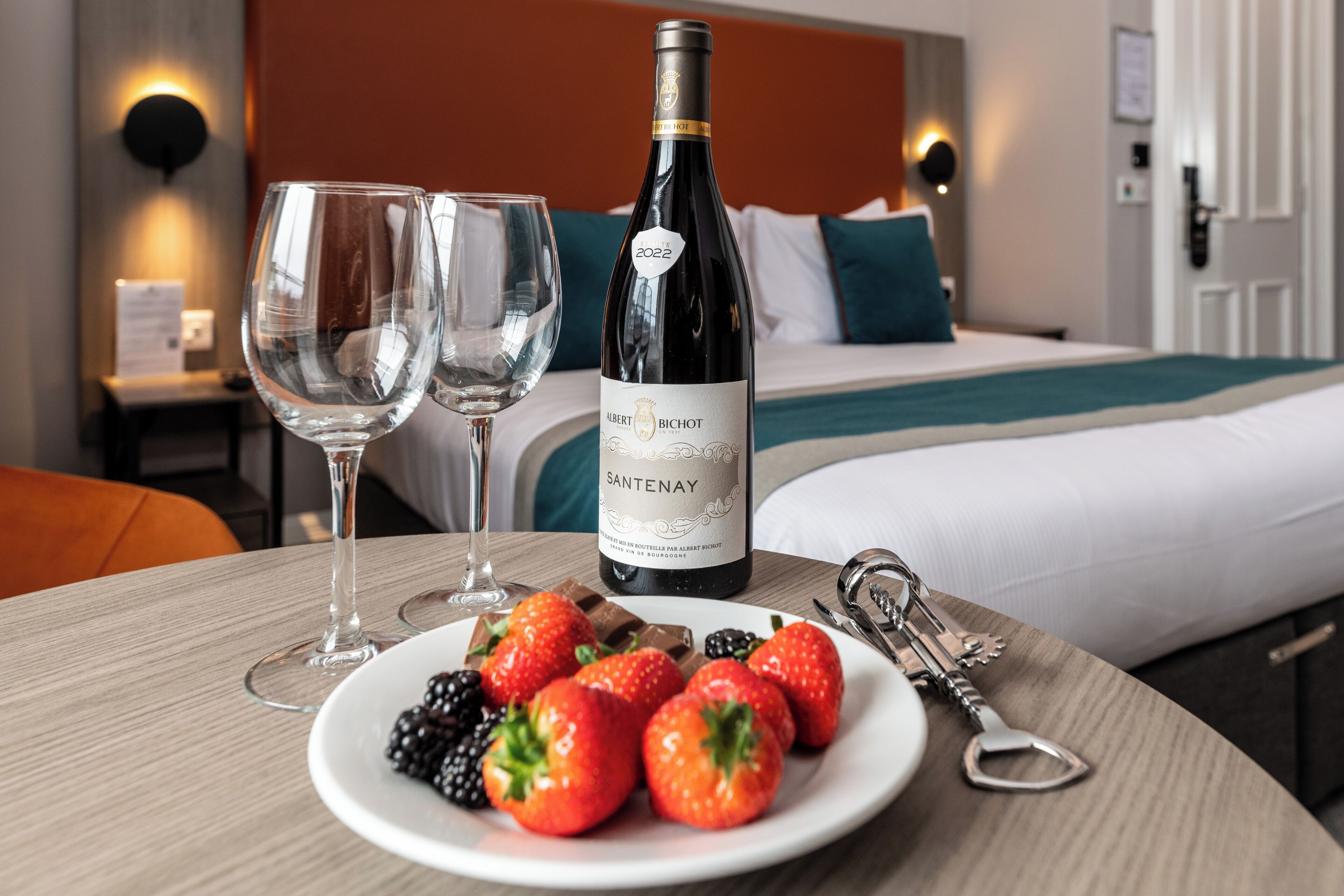A hotel room with a bed, a bedside table with a wine bottle, glasses, and a plate of fruit.