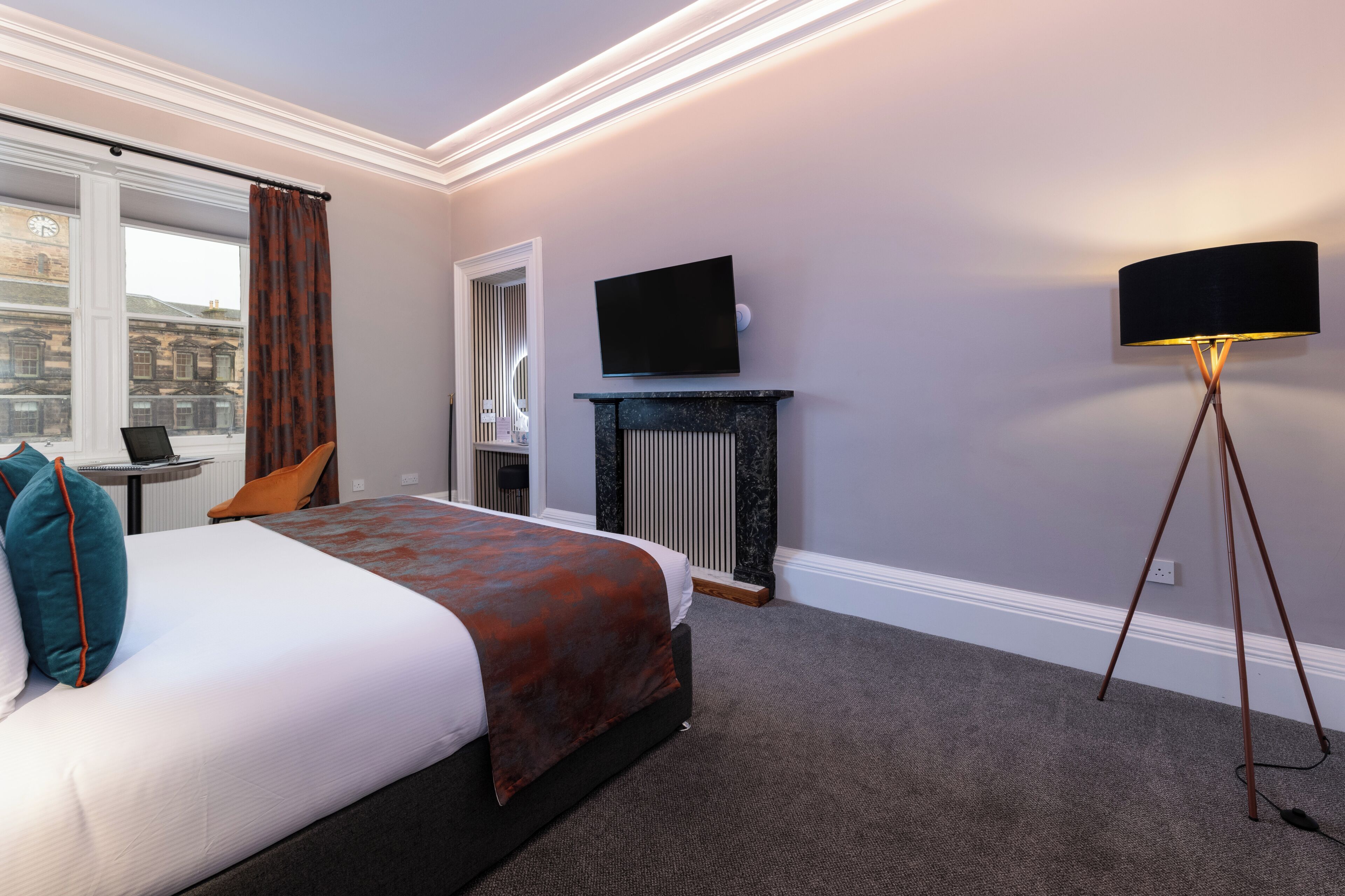 A hotel room with a bed, a television, a standing lamp, a window with curtains, and a view of a building outside.
