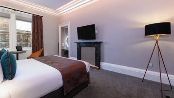 A hotel room with a bed, a television, a standing lamp, a window with curtains, and a view of a building outside.