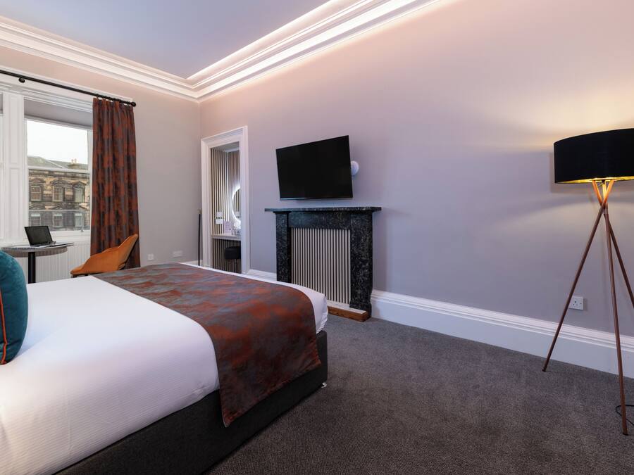 A hotel room with a bed, a television, a standing lamp, a window with curtains, and a view of a building outside.