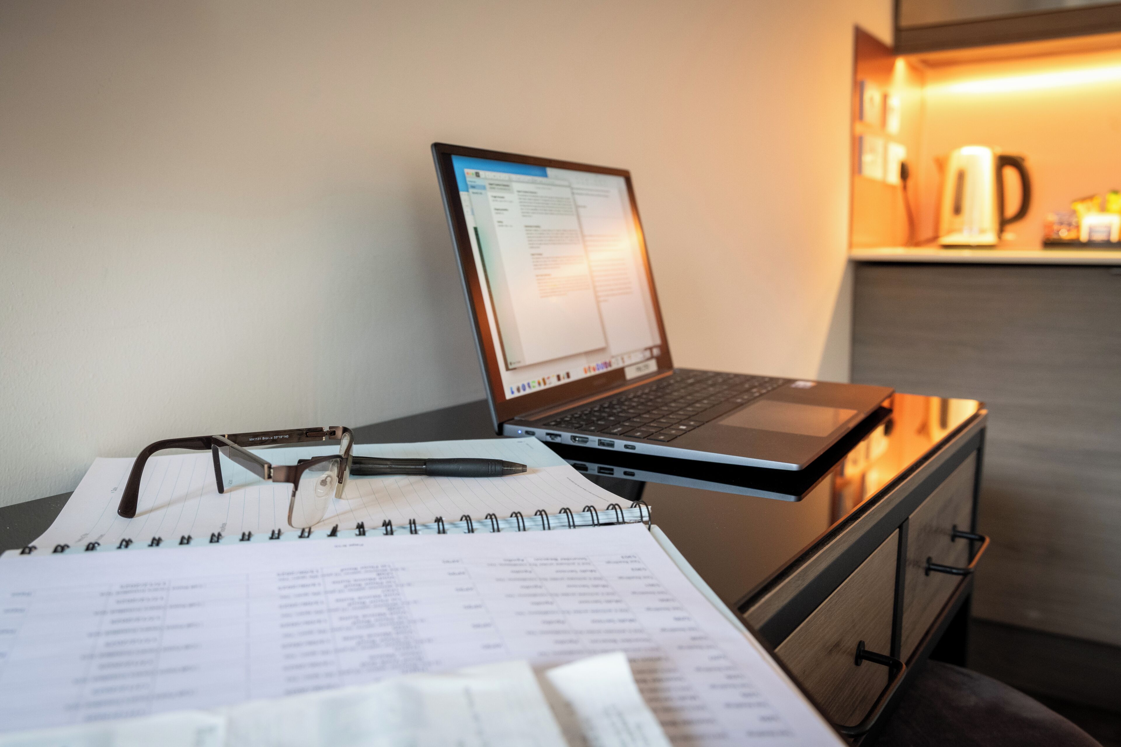 A laptop on a desk with a notebook, glasses, and a pen.
