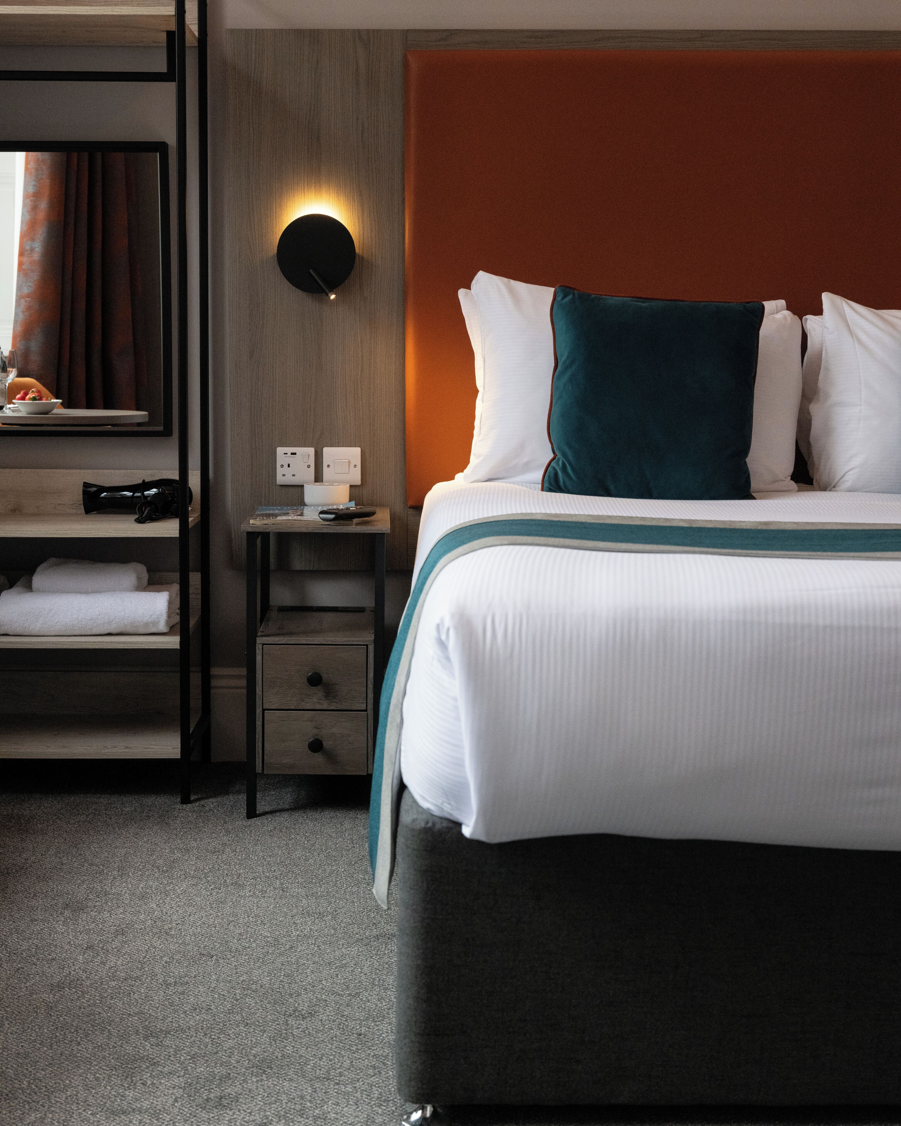 Habitación de hotel con cama, mesita de noche, espejo, luz adosada a la pared y una pequeña estantería con una toalla.