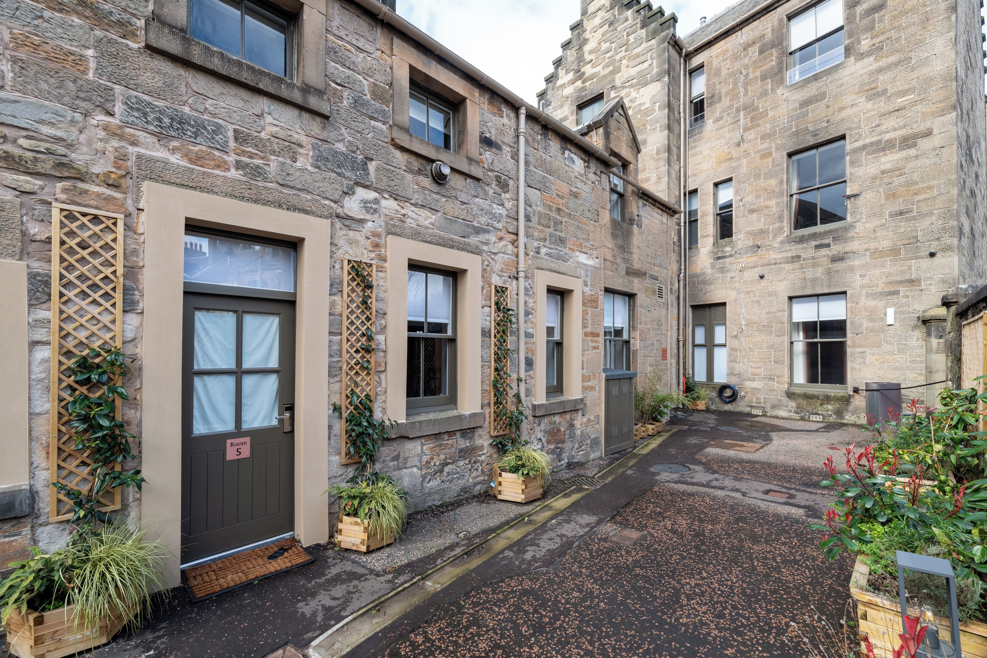 A stone building with multiple windows and doors, some with curtains, and a small garden area with potted plants.