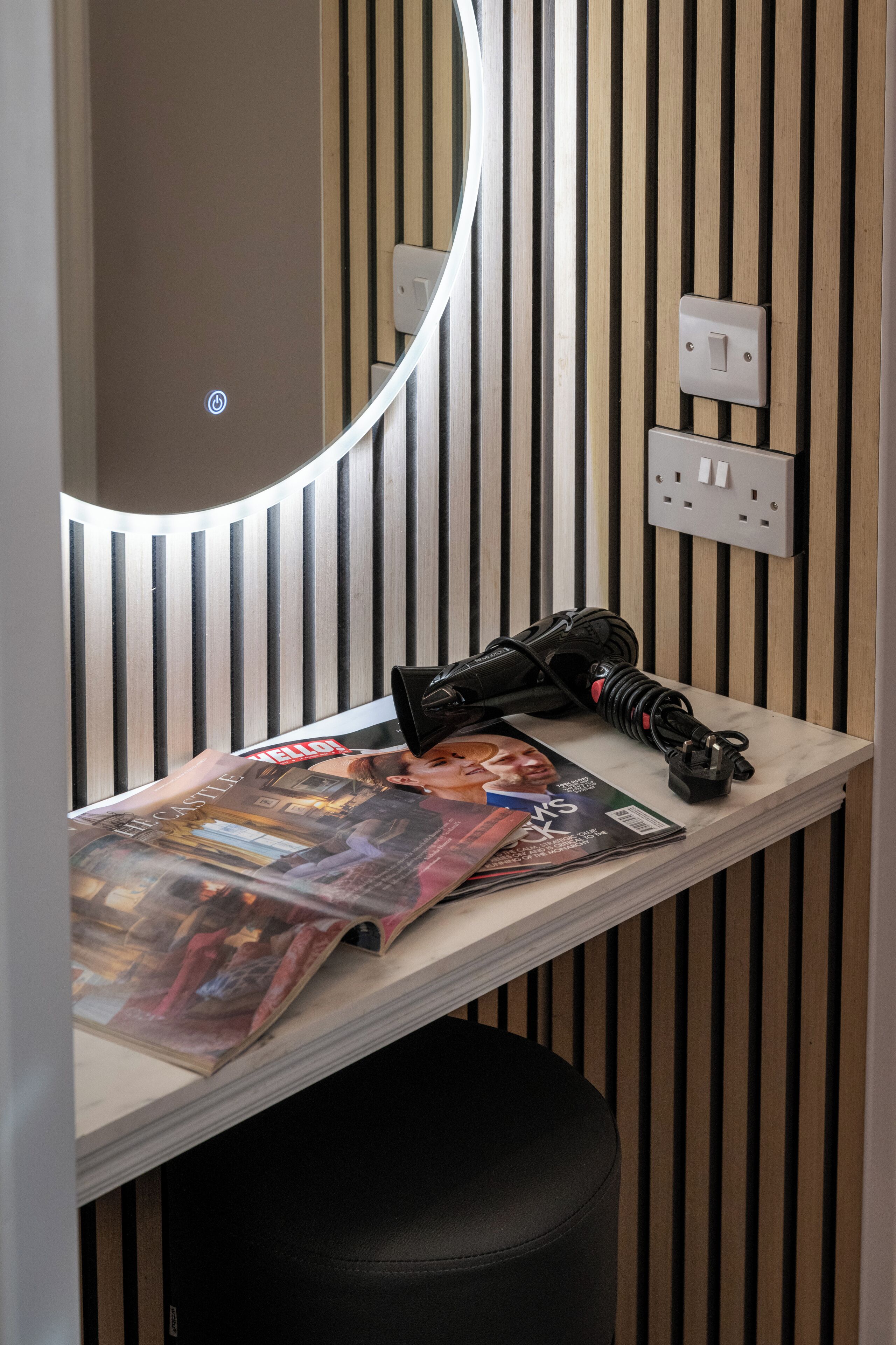 A room with a wooden slat wall, a round mirror, a small table with a magazine, and a black stool.