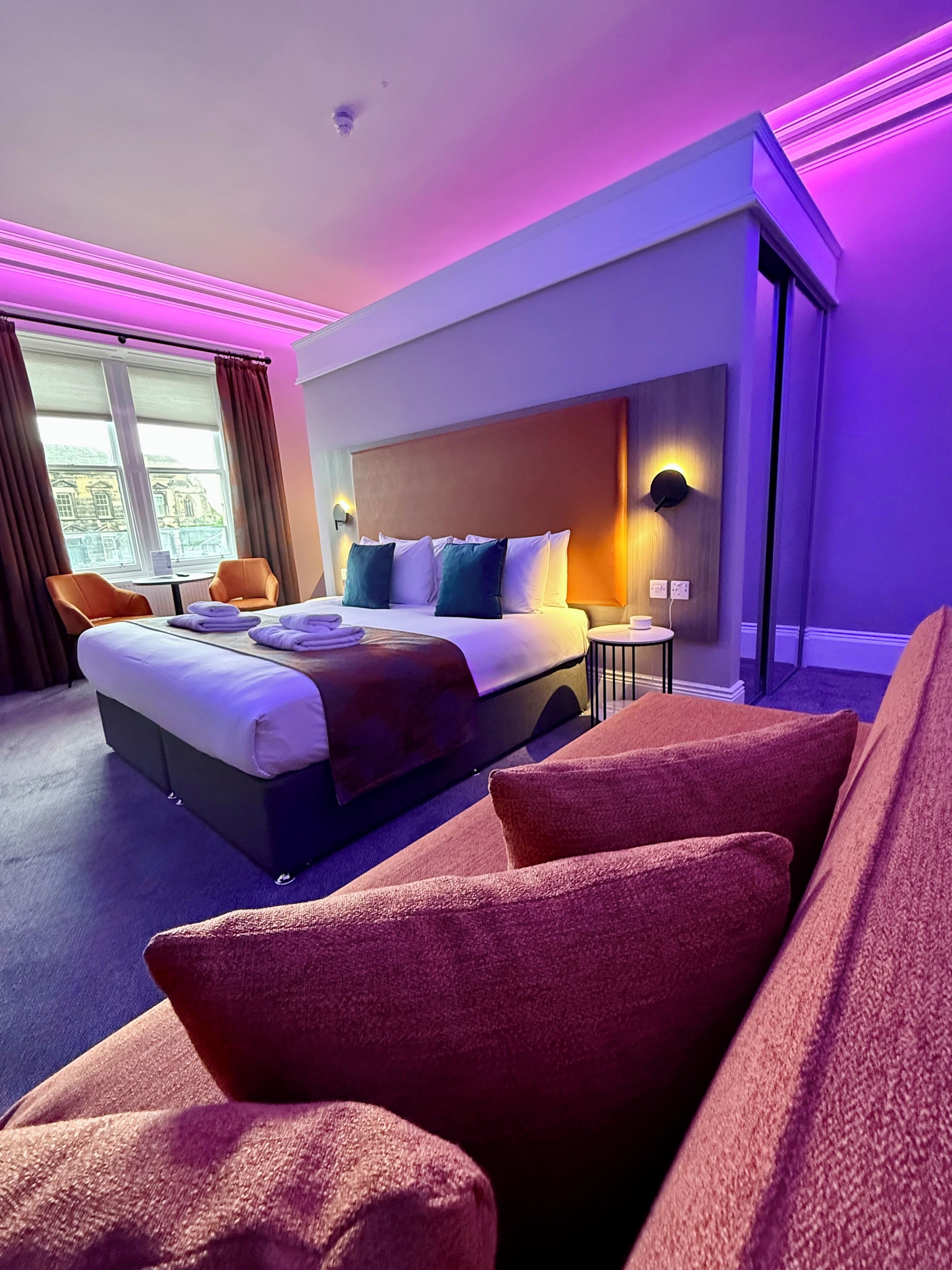 Hotelkamer met een bed, een zithoek met stoelen, een raam met gordijnen en uitzicht op een gebouw.