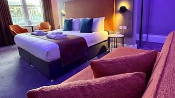 Habitación de hotel con cama, zona de estar con sillas, ventana con cortinas y vista a un edificio.