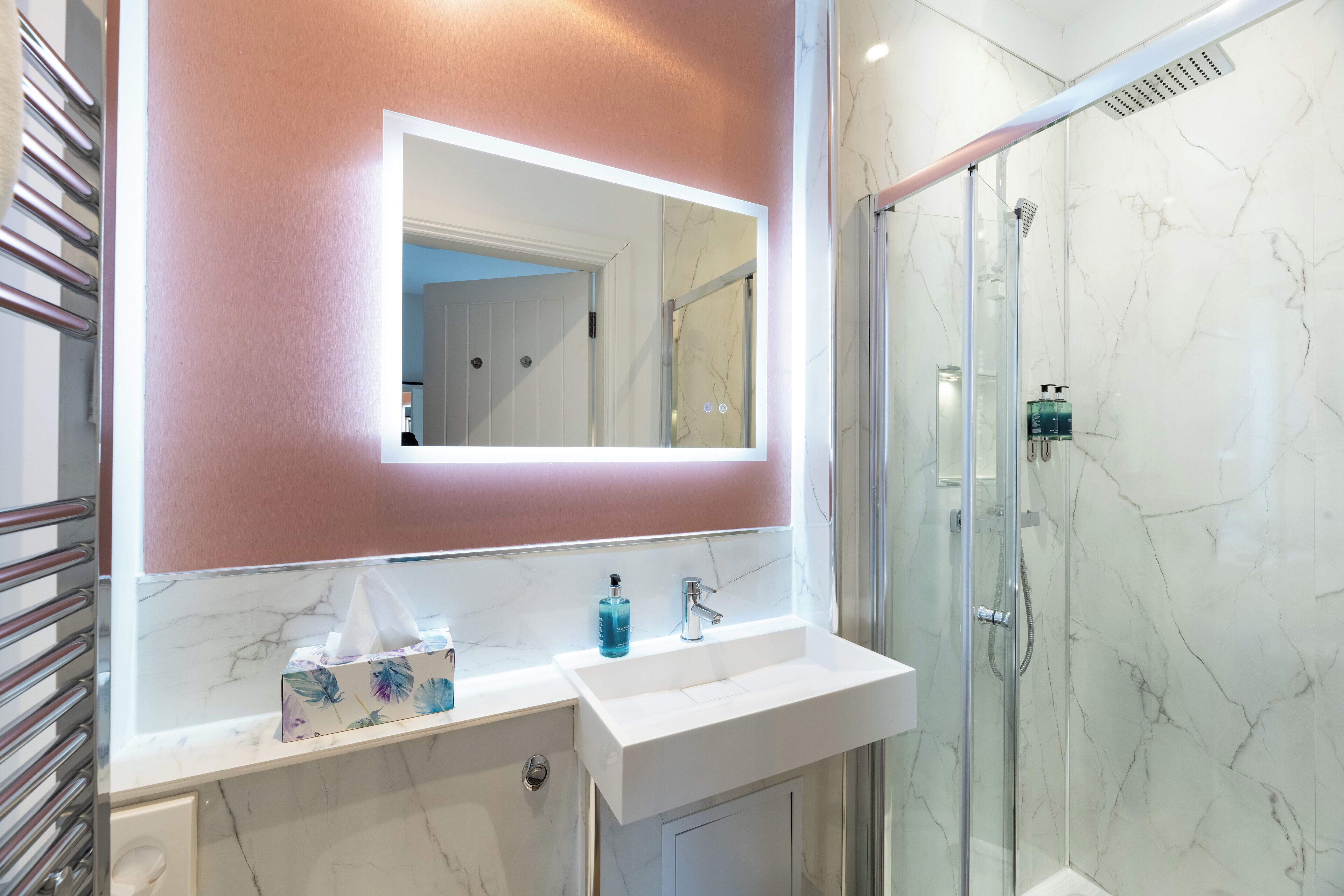 Un baño con un lavabo blanco, cabina de ducha con mampara de vidrio y espejo sobre una pared rosa.