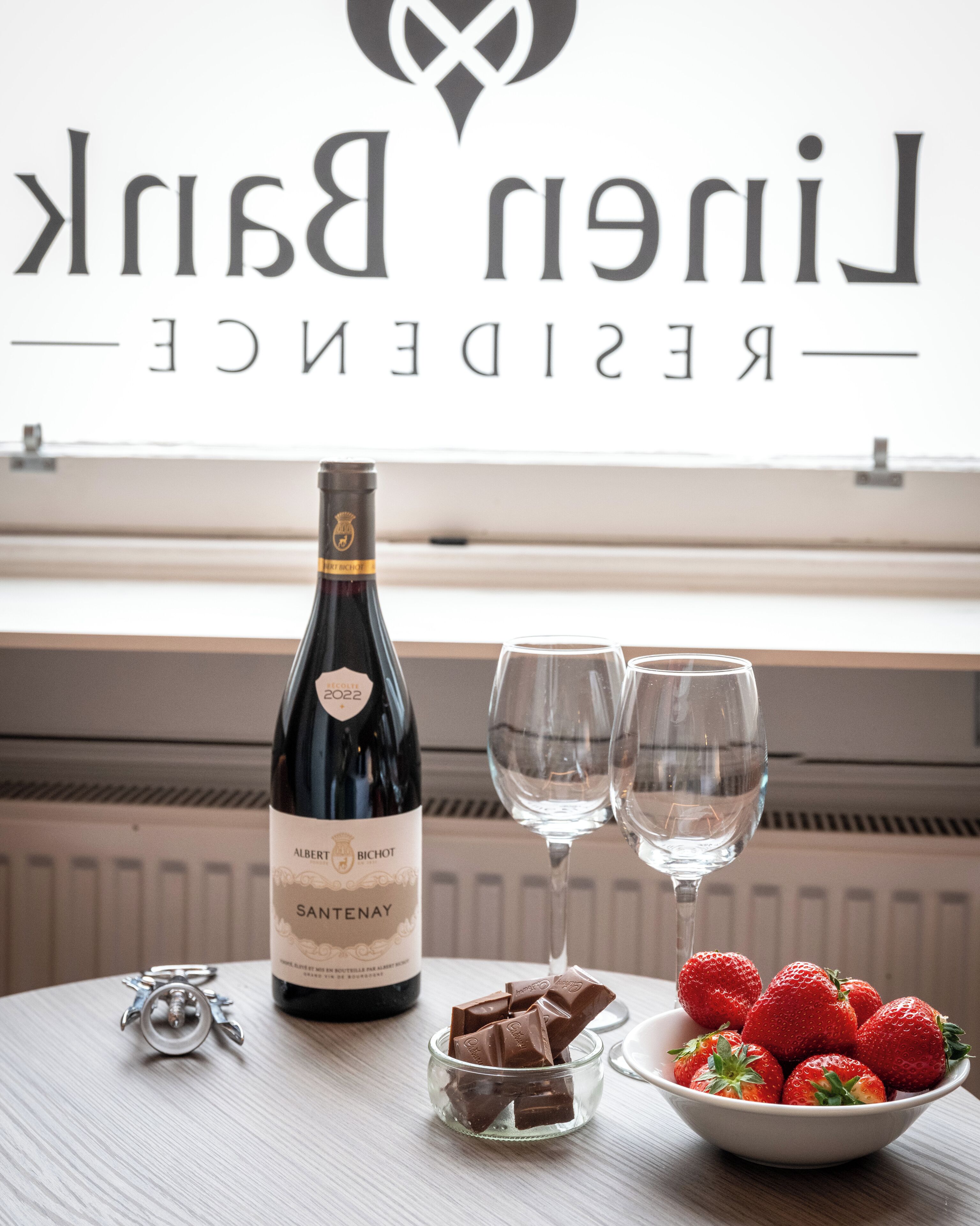 Una botella de vino, dos copas y un cuenco con fresas sobre una mesa, con un cartel de fondo.