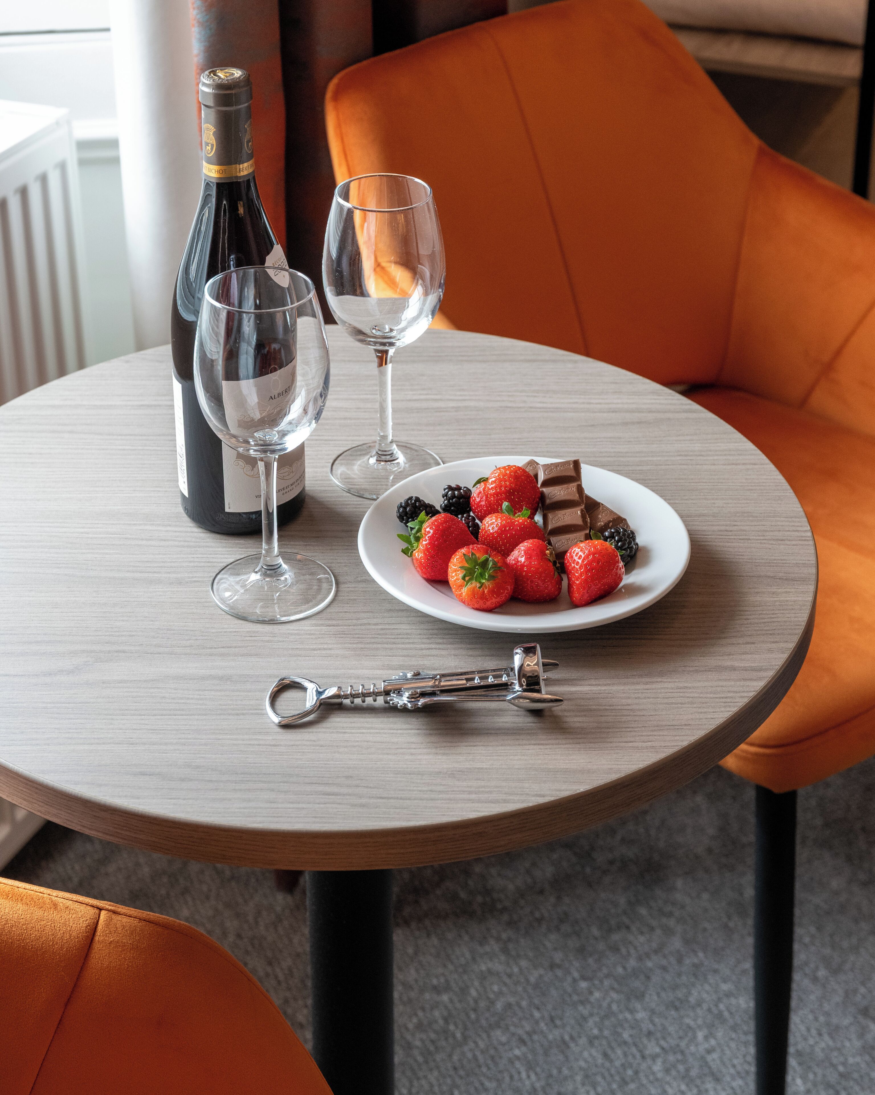 A round table with a bottle of wine, two wine glasses, a plate of strawberries and blackberries, and a bottle opener.