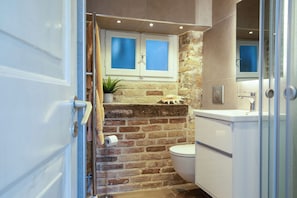 Une salle de bain avec un mur en briques, des toilettes, un lavabo et une fenêtre située au-dessus des toilettes.