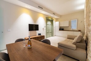 A modern living room with a wooden dining table, a beige sofa, a TV mounted on the wall, and a kitchen area in the background.