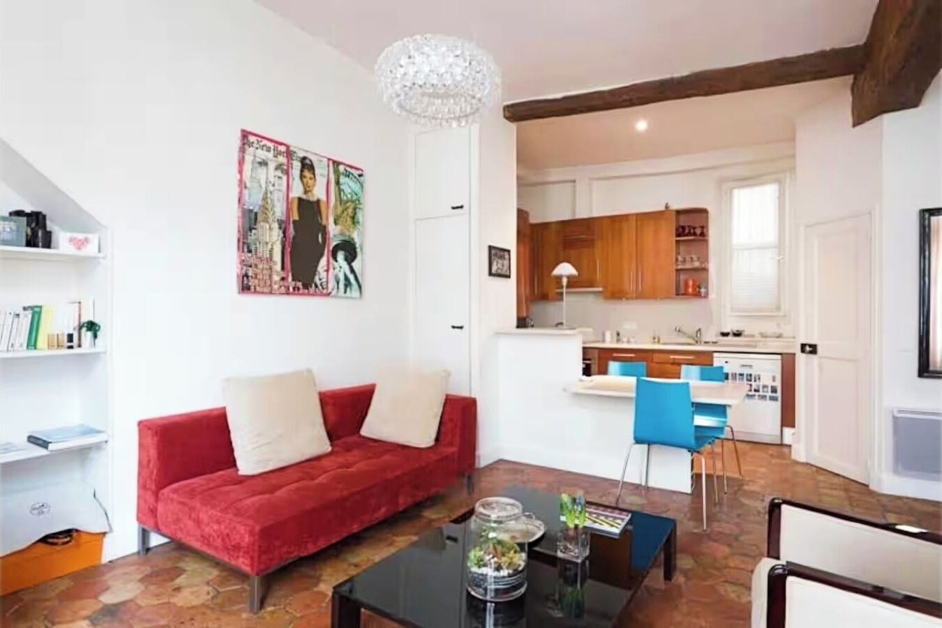 A living room with a red sofa, a coffee table, and a dining area with blue chairs. There is a kitchen visible in the background.