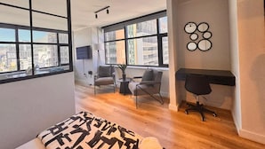 Een modern appartement met grote ramen, een bureau, twee stoelen en een bed met beddengoed met een patroon.