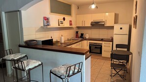 A compact kitchen with a central island, a sink, a stove, and a refrigerator. There are two bar stools and a small dining table.