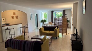 A modern living room with a sofa, a coffee table, a dining table with chairs, a kitchen area with a counter and stools, and large windows with curtains.