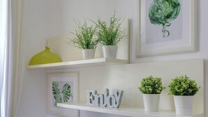 A wall with three white shelves, each holding different items: a yellow vase, a framed plant, and a framed artwork.