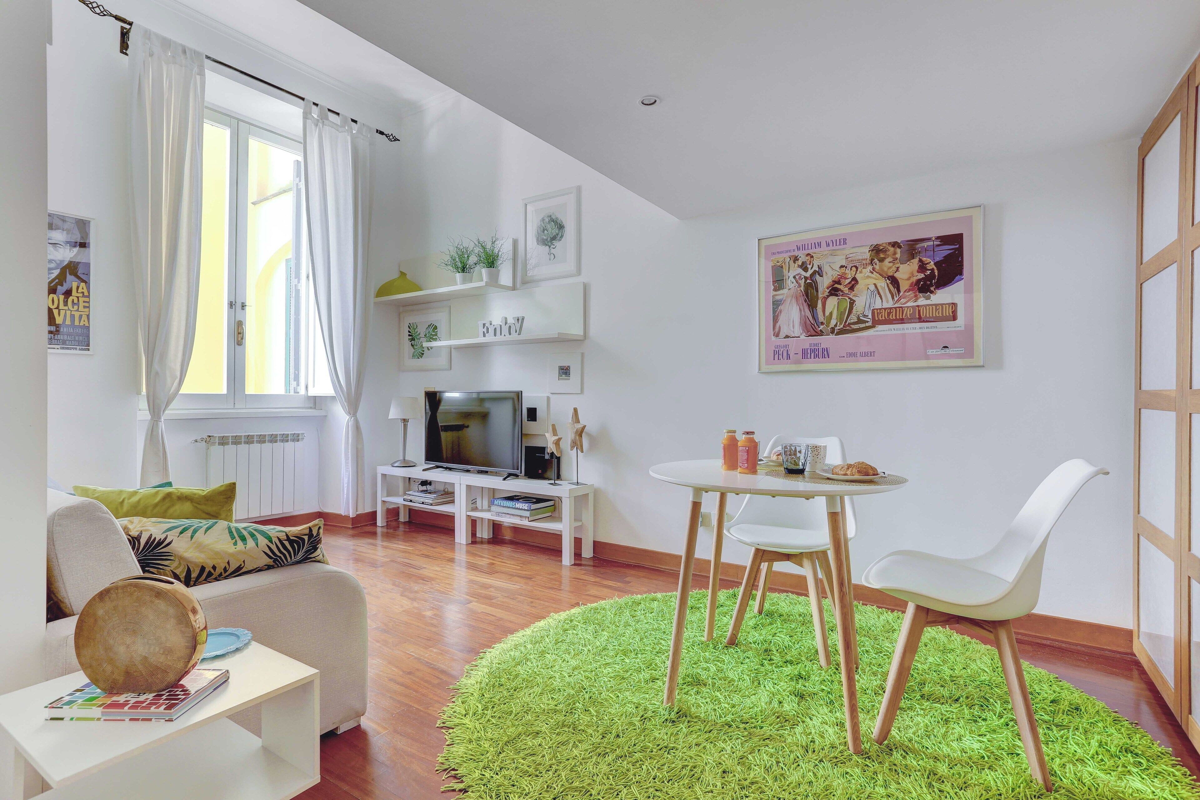 Un salon moderne doté d’un téléviseur à écran plat, d’un coin repas avec une petite table et des chaises, et d’un tapis à poils longs de couleur verte.