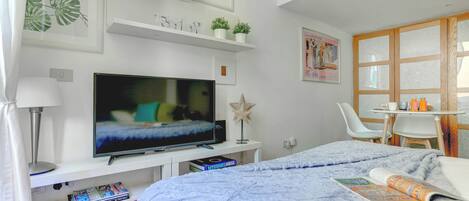Un dormitorio moderno con una gran televisión de pantalla plana, una cama con una manta azul, una mesita de noche con una taza y un libro, y una silla.