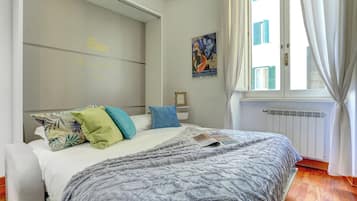 Quarto com cama, travesseiros, janela com cortinas, radiador e um pôster na parede.