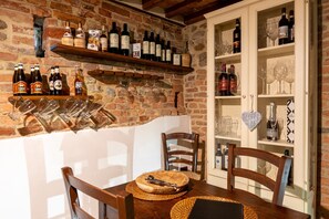 A dining area with a brick wall, wooden chairs, a table with a placemat, wine bottles on shelves, and wine glasses.