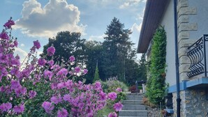 Un bâtiment en pierre avec un escalier menant à une porte, le tout entouré d’une végétation luxuriante et de fleurs violettes.