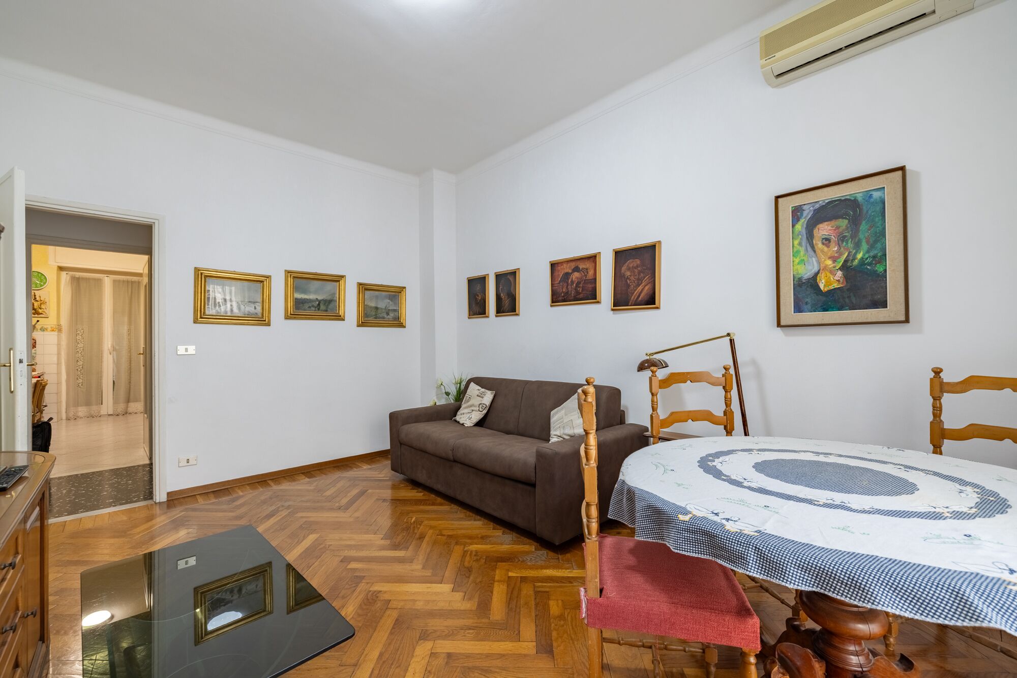 Una sala de estar con un sofá marrón, una mesa de comedor de madera con mantel a cuadros, y varias imágenes enmarcadas en la pared.