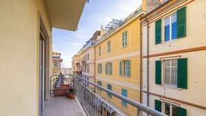 Un balcon étroit doté d’une rambarde en métal, de plantes en pot et offrant une vue sur les bâtiments voisins, qui arborent des volets verts.
