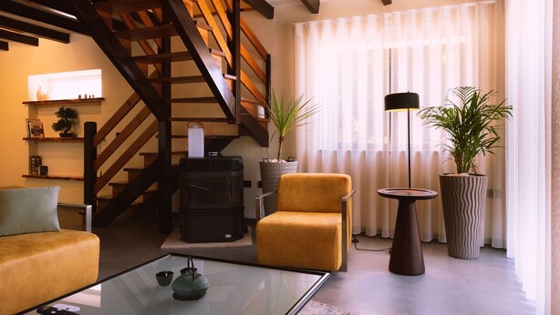 Una sala de estar con un sofá, una mesa de centro de vidrio, una chimenea, una escalera y ventanas amplias con cortinas ligeras.